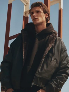 Model wearing a dark shearling-lined leather bomber jacket and black hoodie against an outdoor red and white structure