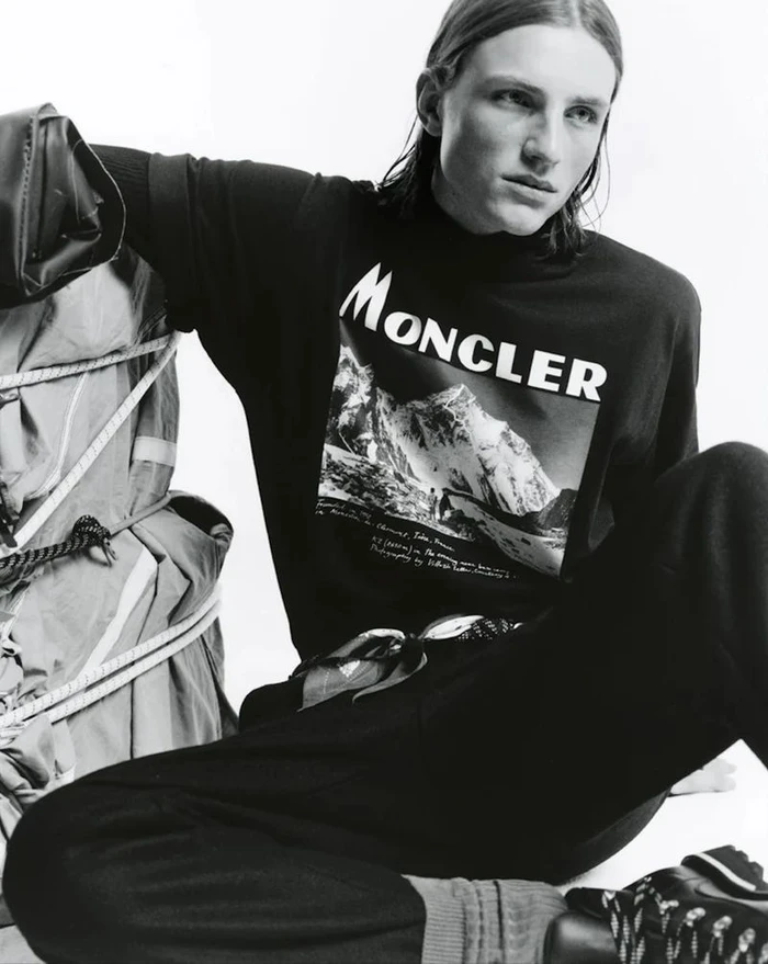Model wearing a black Moncler sweatshirt with a mountain graphic seated against a plain white background
