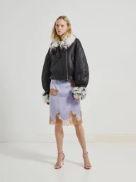 Model wearing a black shearling-trimmed jacket and lavender slip skirt against a seamless white studio background