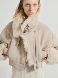 Model wearing a cream shearling aviator jacket with a wide collar and fluffy cuffs against a light background
