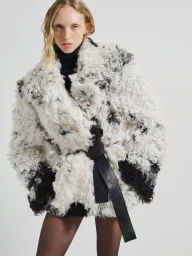 Model wearing a textured white and black patterned shearling coat with a black leather belt against a neutral background.