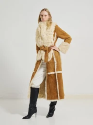 Model in long brown shearling coat wearing black boots against a plain white studio background