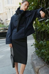 Model leaning against a dark metal fence wearing a navy jacket and black skirt holding a black handbag on a city street.