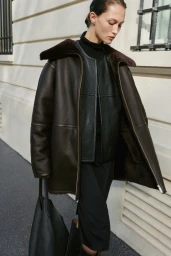 Model wearing a dark shearling-lined coat and black leather top walking past a classical building exterior.