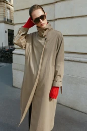 Model wearing dark sunglasses, a beige trench coat, and bright red knit sleeves outdoors against a stone building.