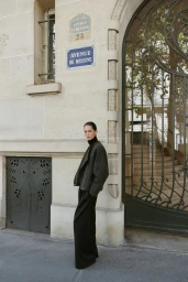 Model Vika Evseeva standing outdoors on a sidewalk against a classical stone building facade with large iron gates.