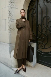 Model wearing a brown coat and trousers holding a dark handbag in front of an arched doorway and stone wall