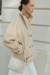 Model wearing a beige double-breasted collared jacket and holding a cream handbag against a cream building exterior wall.