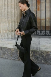 Model Vika Evseeva wearing a black leather jacket and trousers holding a handbag against a stone building exterior