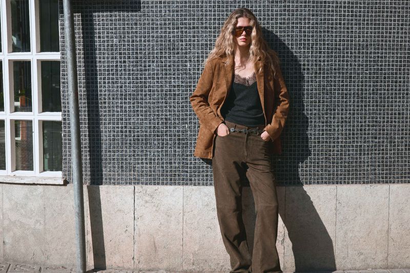 Model wearing a brown jacket and trousers posing against a dark tiled wall outdoors in natural light.