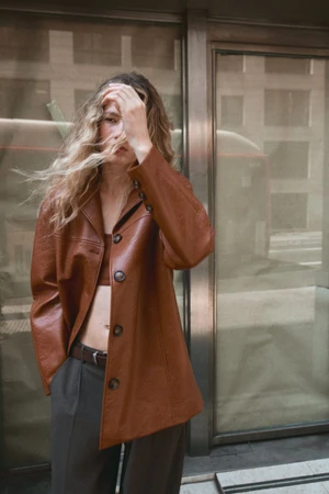 Model with blonde hair wearing a brown leather jacket and dark trousers standing against an urban glass storefront