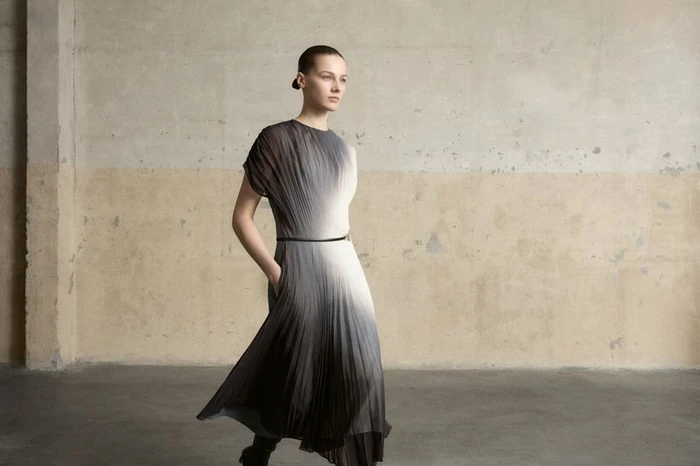 Model wearing a pleated ombré dress with a thin belt standing against a weathered concrete wall.