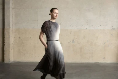 Model wearing a pleated ombré dress with a thin belt standing against a weathered concrete wall.