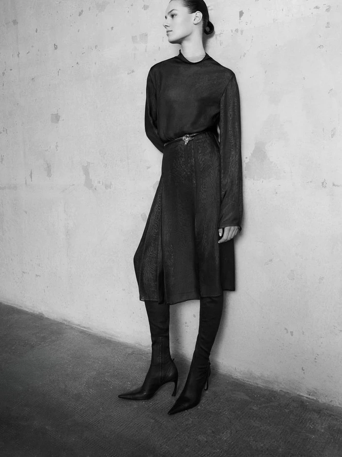 Model Famke van Hasselt wearing a dark belted dress and boots against a textured concrete wall for Giada FW26