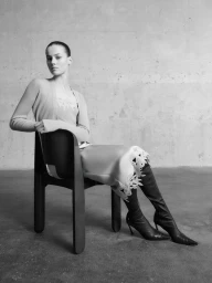 Model Famke van Hasselt seated on a dark chair against a concrete wall for GIADA FW26 Pre Fall Collection