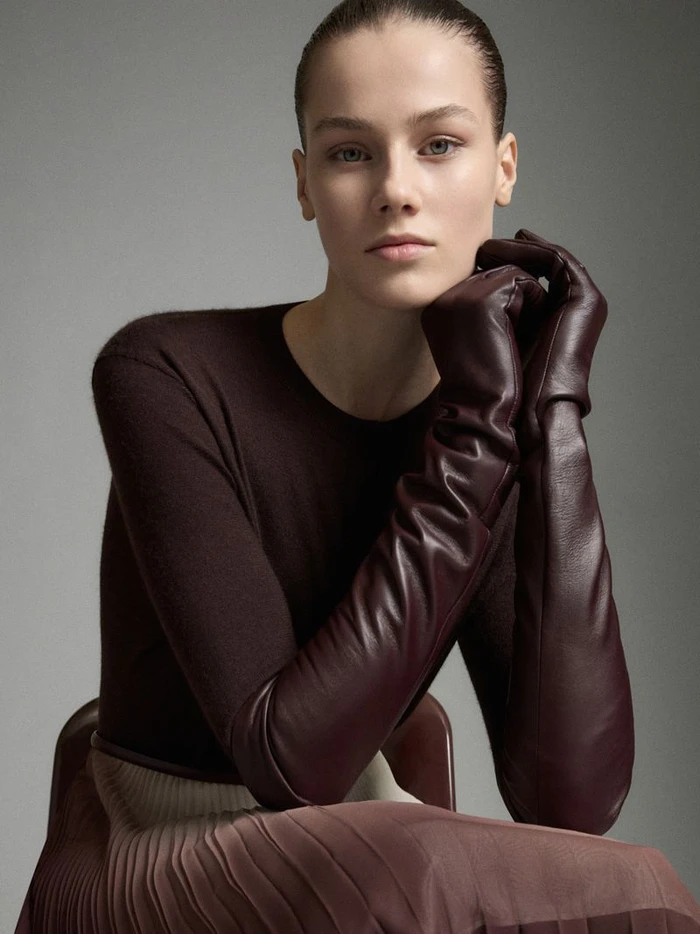 Model wearing dark burgundy leather opera gloves and dark sweater sitting against a neutral grey studio backdrop