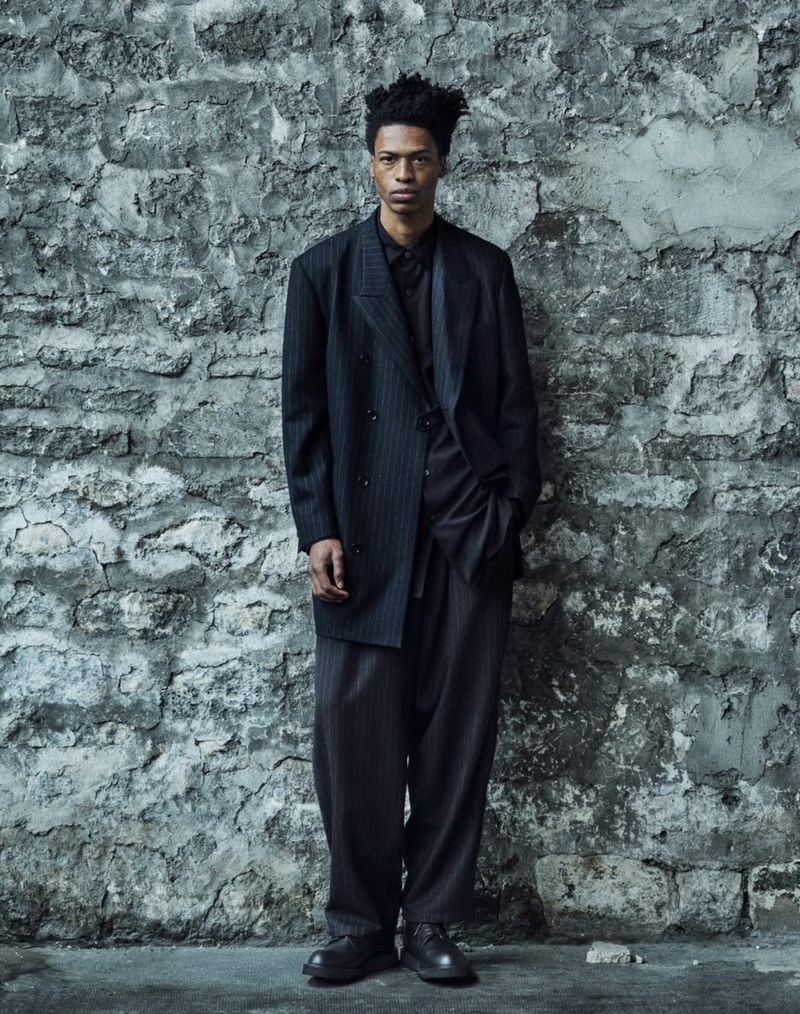 Man in a pinstripe suit poses against a weathered stone wall, showcasing fashion and style in an indoor studio setting with neutral tones.