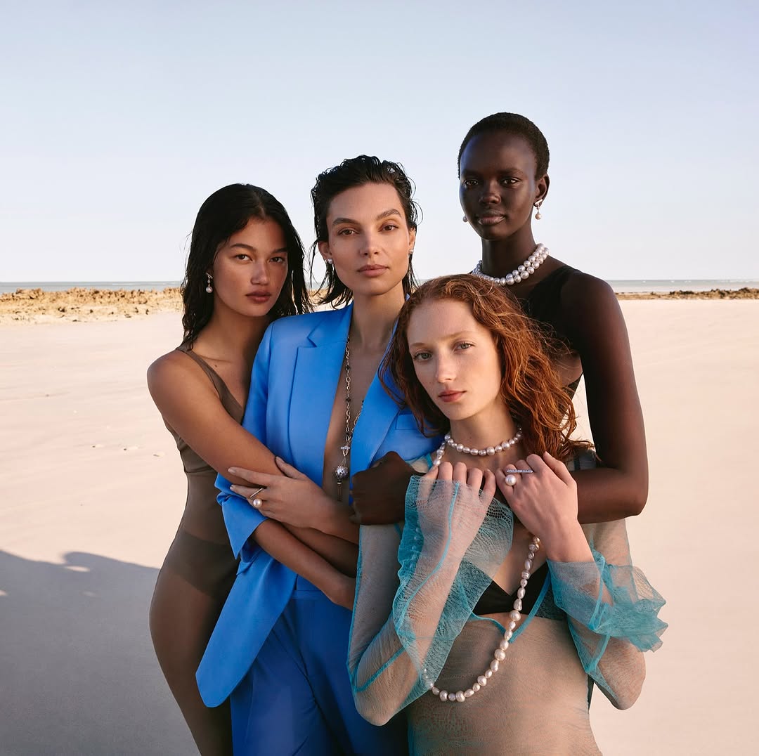 Four diverse female models pose on a beach wearing pearl necklaces, earrings, and rings, showcasing fashion and beauty in natural light.