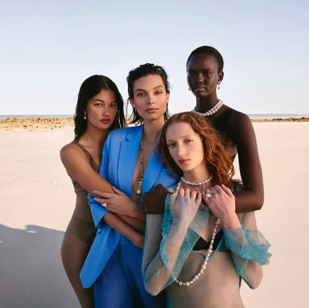 Four diverse female models pose on a beach wearing pearl necklaces, earrings, and rings, showcasing fashion and beauty in natural light.