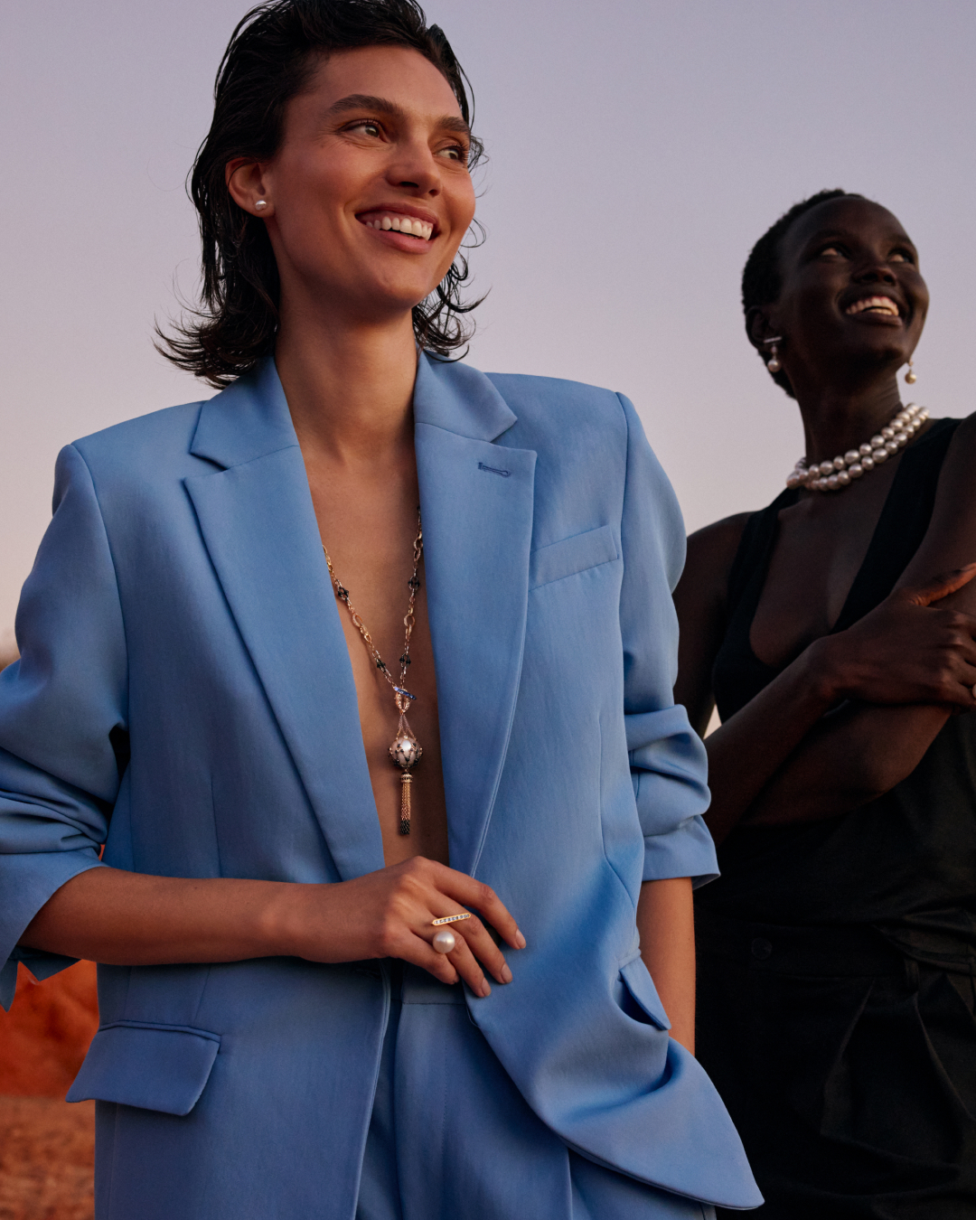 Two women in blazers and jewellery smile in an outdoor setting, against a pale horizon with warm earth tones. Fashion shot.