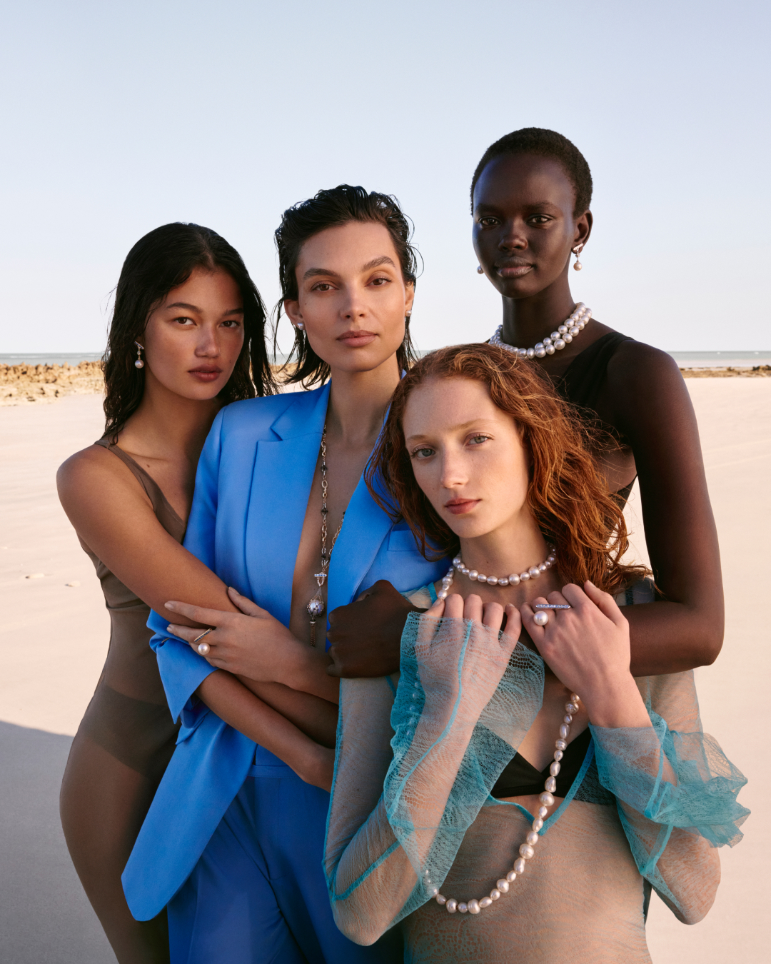 Four women of diverse ethnicities model pearl jewelry on a light sandy beach. The focus is on the pearl necklaces, earrings and rings.