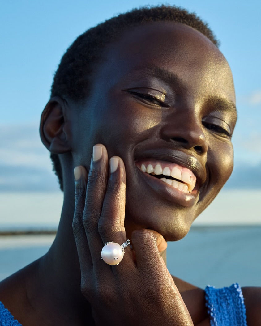 Close-up shot of a smiling dark-skinned woman with natural hair, wearing a pearl ring, posing against a blurred blue and white sky.