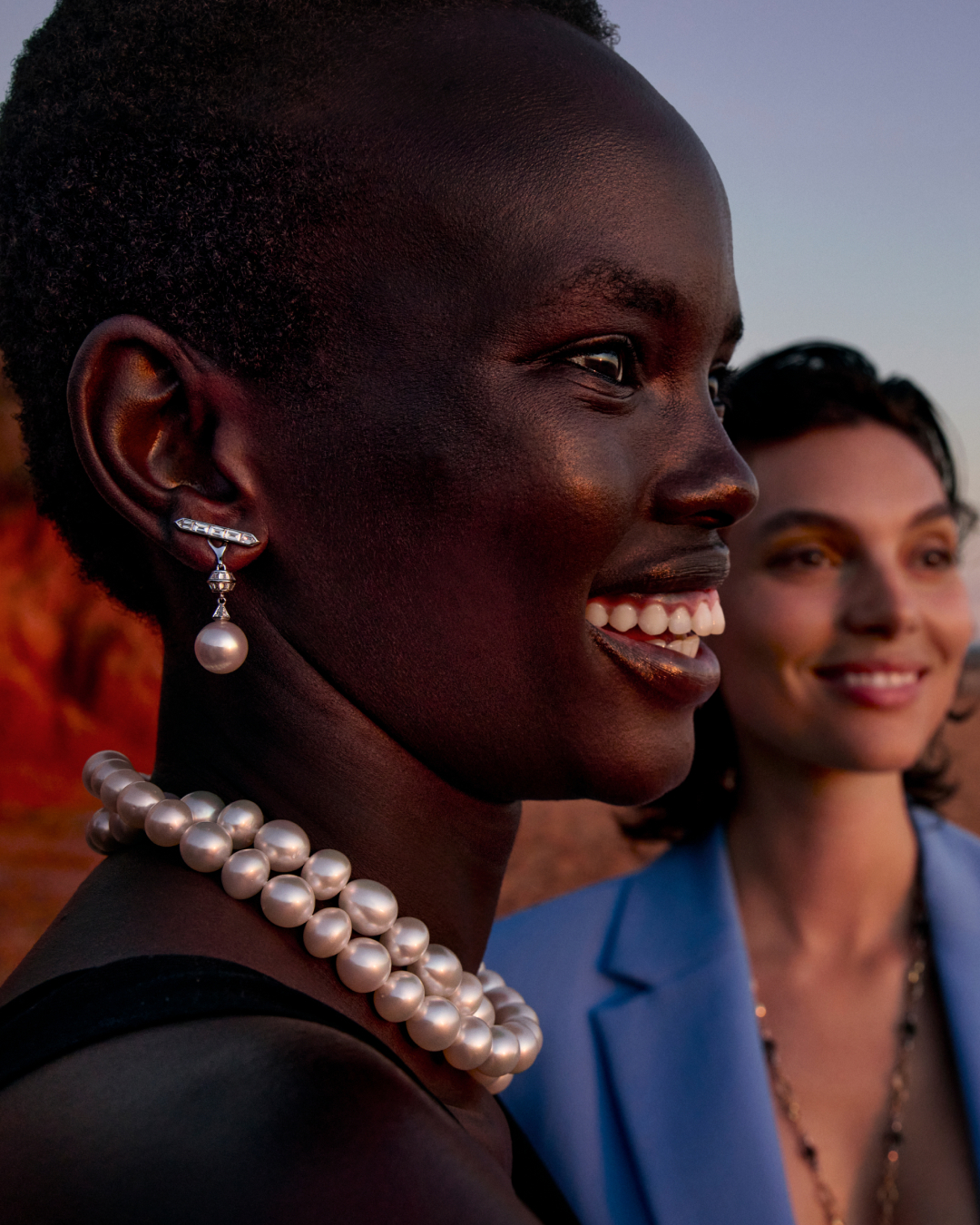 Two women pose outdoors wearing jewelry. The first woman wears a pearl necklace and pearl earring. The second woman wears a blue blazer.