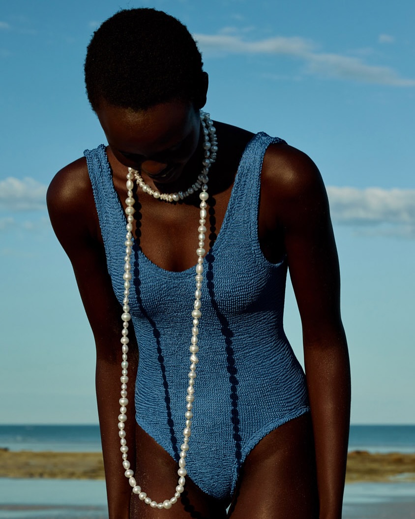 Dark-skinned model in a blue swimsuit stands on a beach with a pearl necklace, under a sunny sky near calm water.