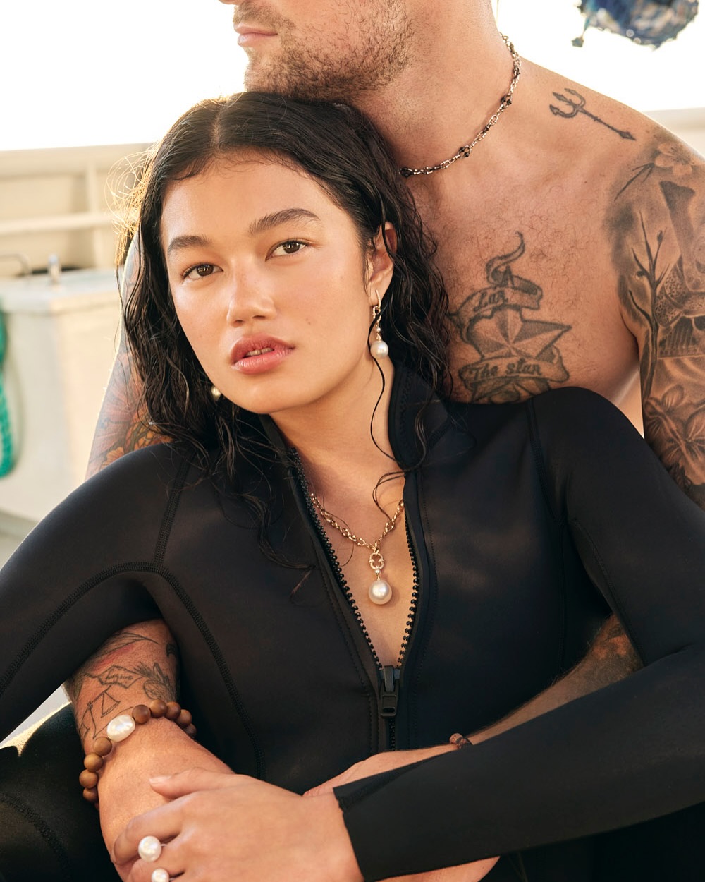 Close-up studio shot of a woman in a black wetsuit and a man with tattoos, both wearing pearl jewelry, under bright, even lighting.
