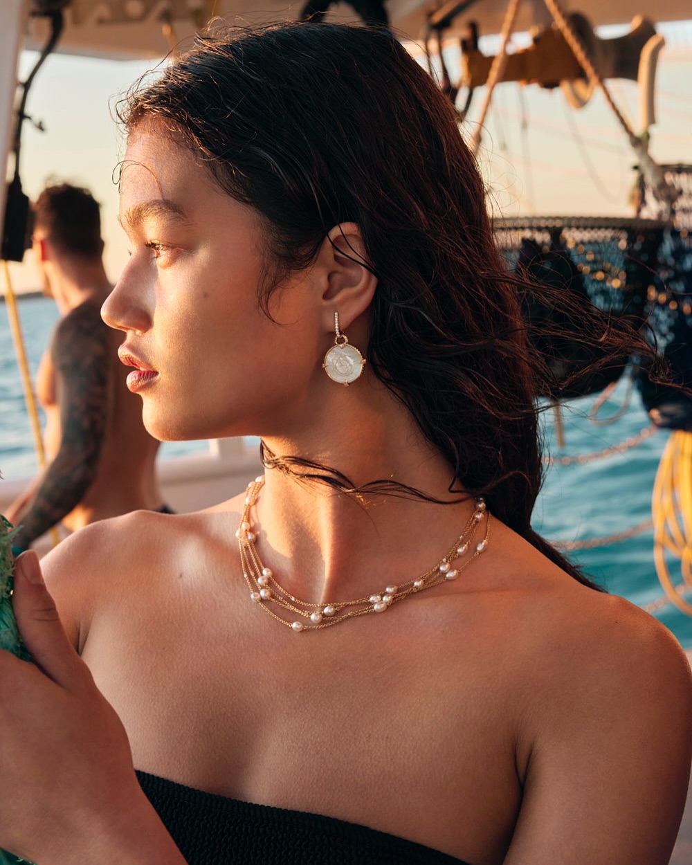 Profile portrait of an adult female wearing a black strapless top, pearl earrings, and layered necklace on a boat with a man in the background.