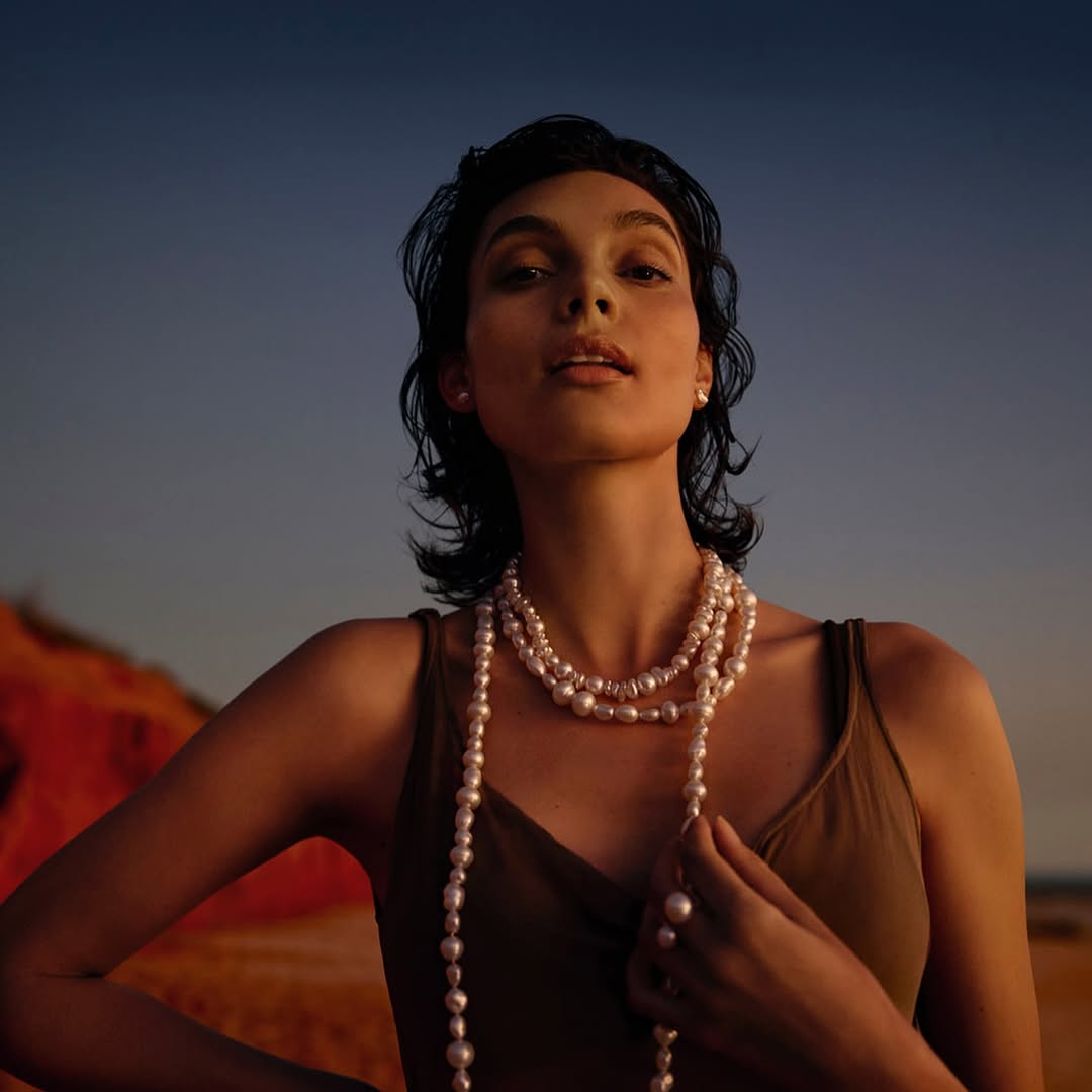 Portrait of a woman with pearl necklaces against a dusk sky, wearing a tank top and natural makeup, showcasing fashion and beauty.