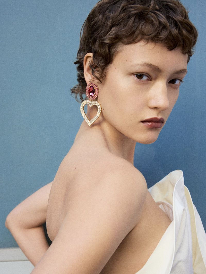 Daria Zolotova wearing a crystal heart drop earring and off-the-shoulder white top against a blue background.