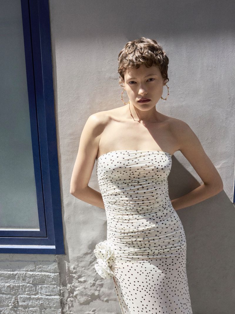 A slender adult woman poses against a concrete wall wearing a strapless, polka-dot dress with her hands behind her back.