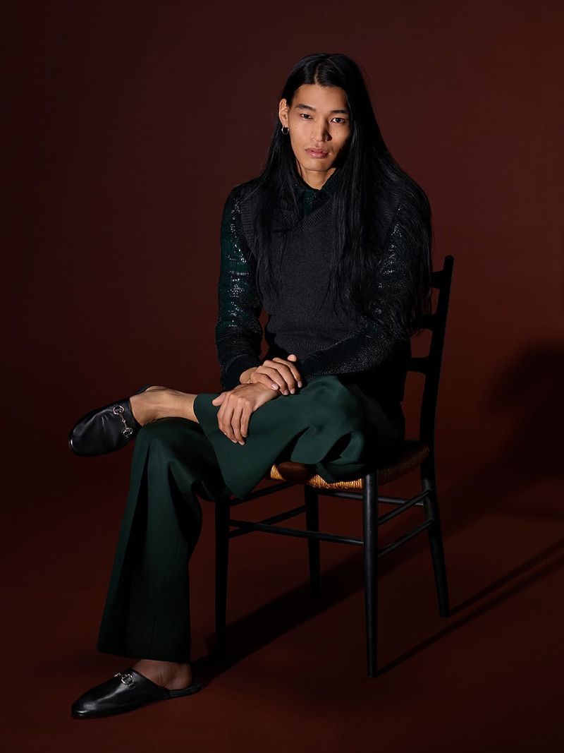 An adult male model with long dark hair is sitting on a chair in a photo studio wearing a shiny long-sleeved top, vest, and dark green trousers.