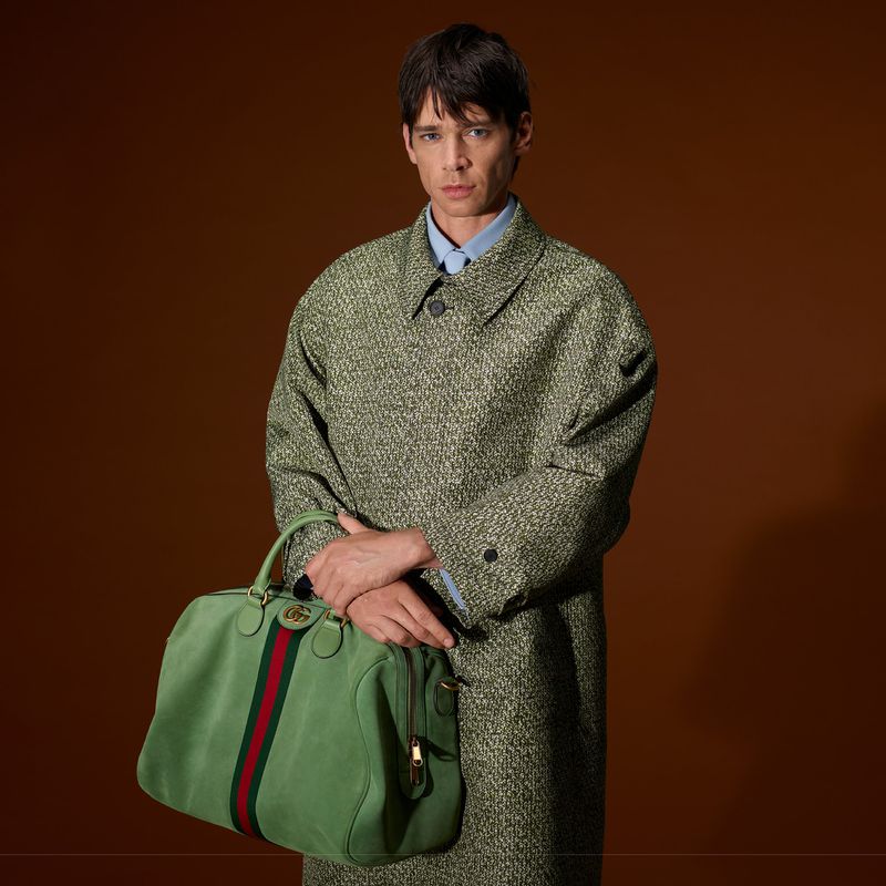 Male fashion model poses in a studio wearing a patterned coat and holding a green Gucci duffel bag against a brown backdrop.