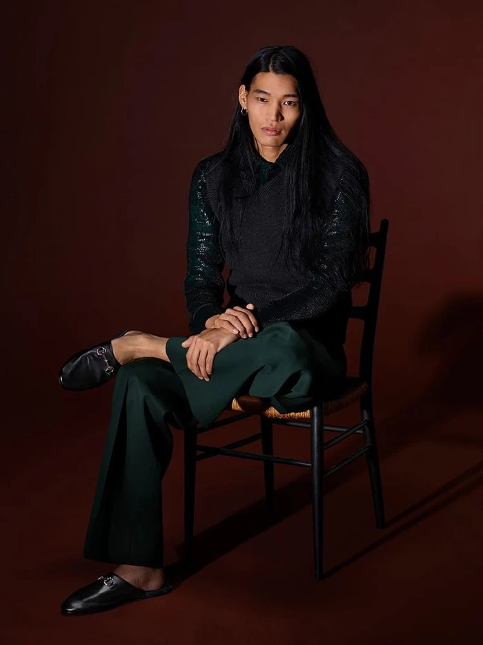 Model with long black hair seated on a wooden chair wearing a dark sweater and green trousers against a dark backdrop