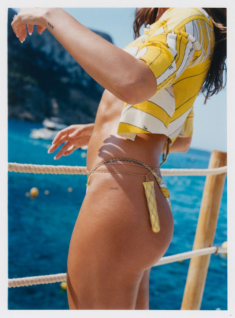 Woman in bikini and patterned crop top poses outdoors with ocean and mountain backdrop, showcasing summer fashion and swimwear.