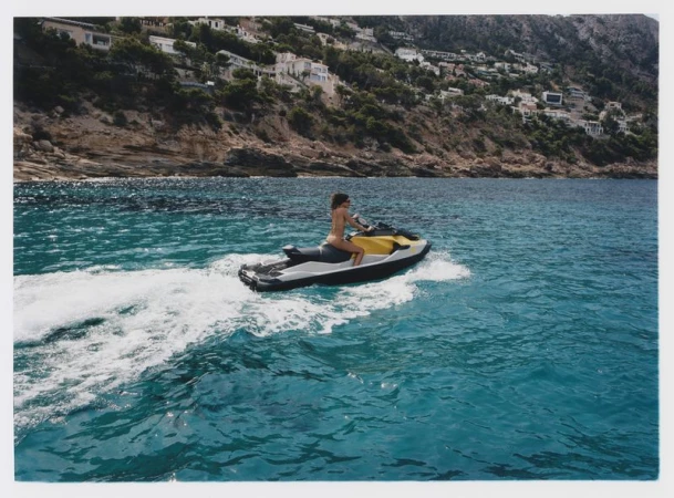 A woman rides a jet ski in the blue ocean near the rocky coast, with cliffside houses dotting the landscape. Sunny day and water sports.