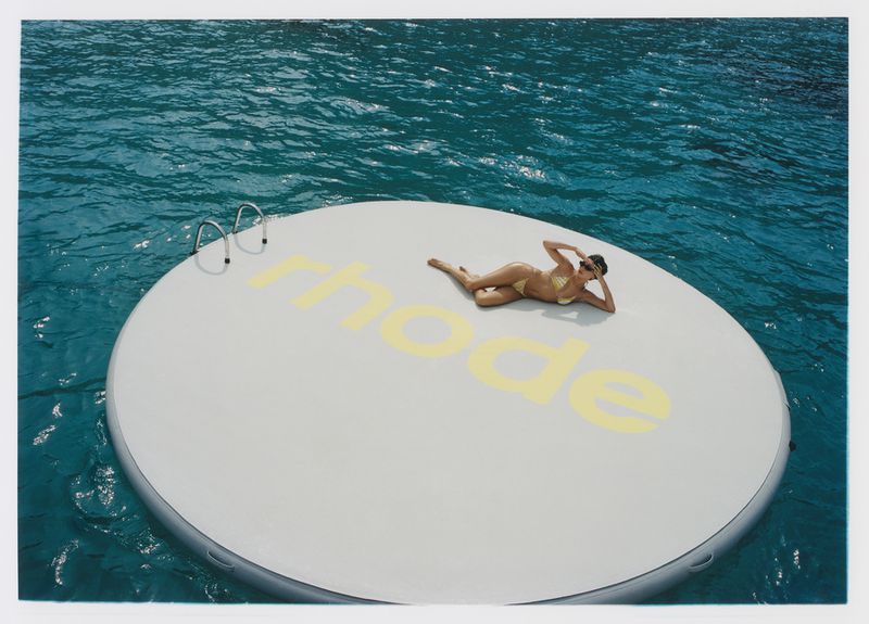 Adult woman in a bikini lies on a floating white platform with a ladder in the clear blue water of the ocean on a sunny day.