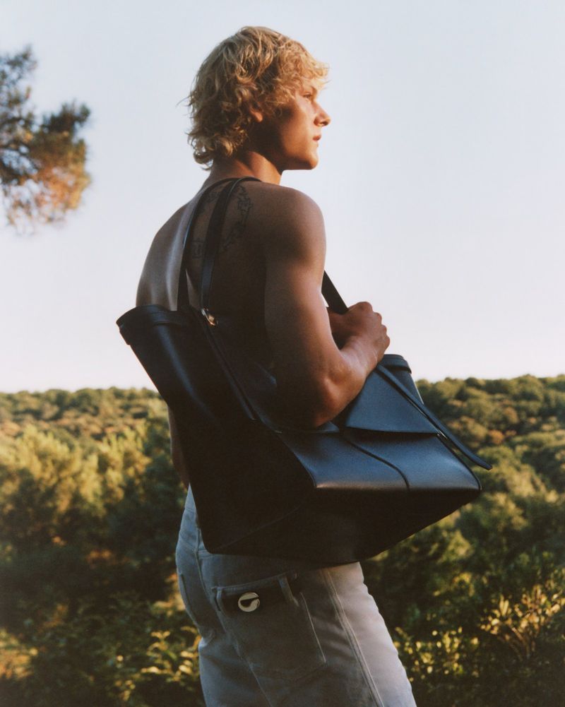 Profile view of a shirtless man with blonde hair, light jeans, and a dark leather tote bag standing outdoors in a nature setting.