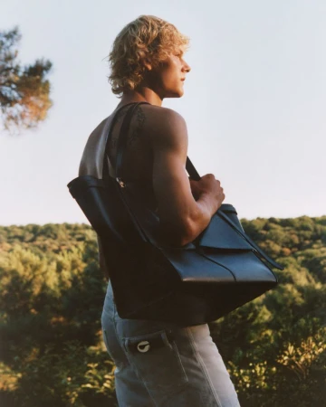 Profile view of a shirtless man with blonde hair, light jeans, and a dark leather tote bag standing outdoors in a nature setting.
