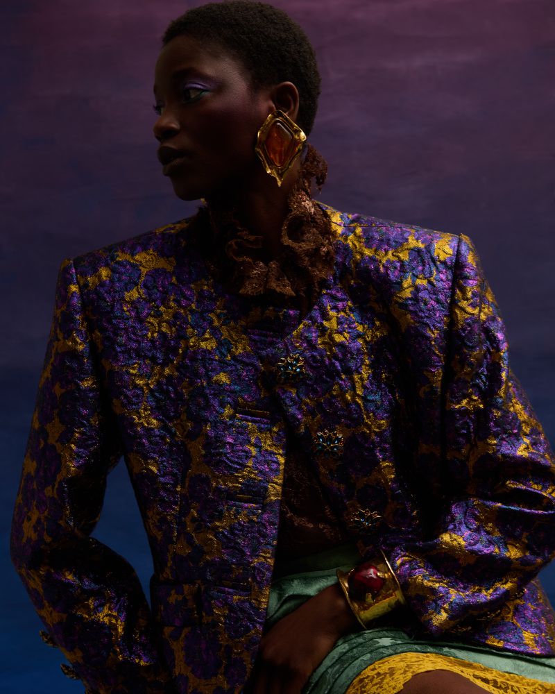Black woman wearing a purple and gold patterned jacket, oversized amber earrings, and a gold cuff. Fashion editorial portrait.