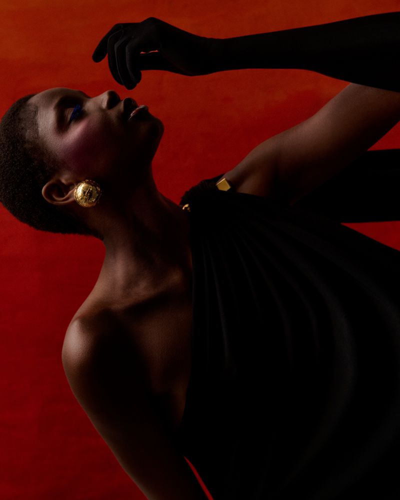 Elegant portrait of a Black woman with short hair wearing a draped black dress and gold earrings against a red backdrop in a fashion editorial.