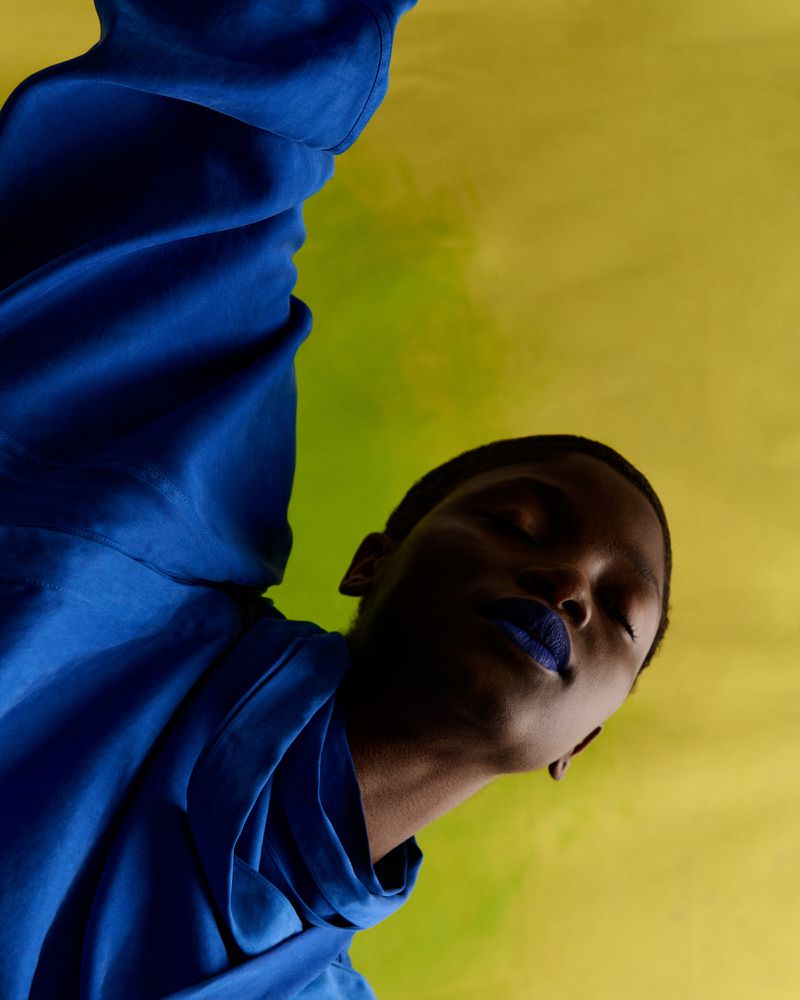 Close-up studio shot of a dark-skinned adult female model with a closed eyes, wearing a draped bright blue top and matching blue lipstick.