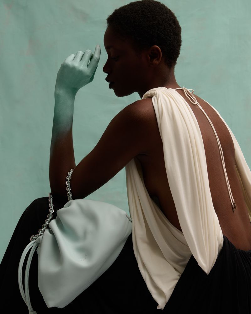 Elegant studio portrait of an African woman with a draped top and light blue handbag, striking a contemplative pose against a blue backdrop.