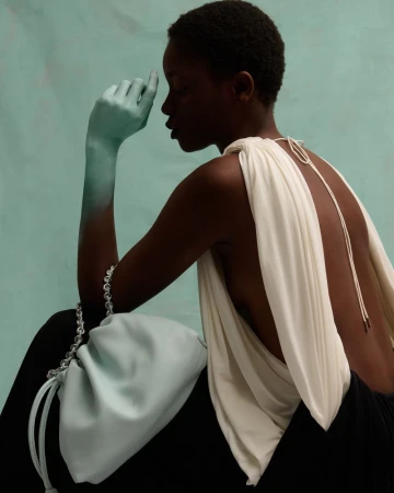 Elegant studio portrait of an African woman with a draped top and light blue handbag, striking a contemplative pose against a blue backdrop.