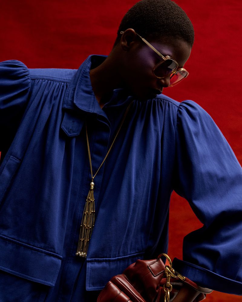 Adult fashion model poses in a blue collared jacket with sunglasses and a gold necklace, against a solid red studio backdrop.