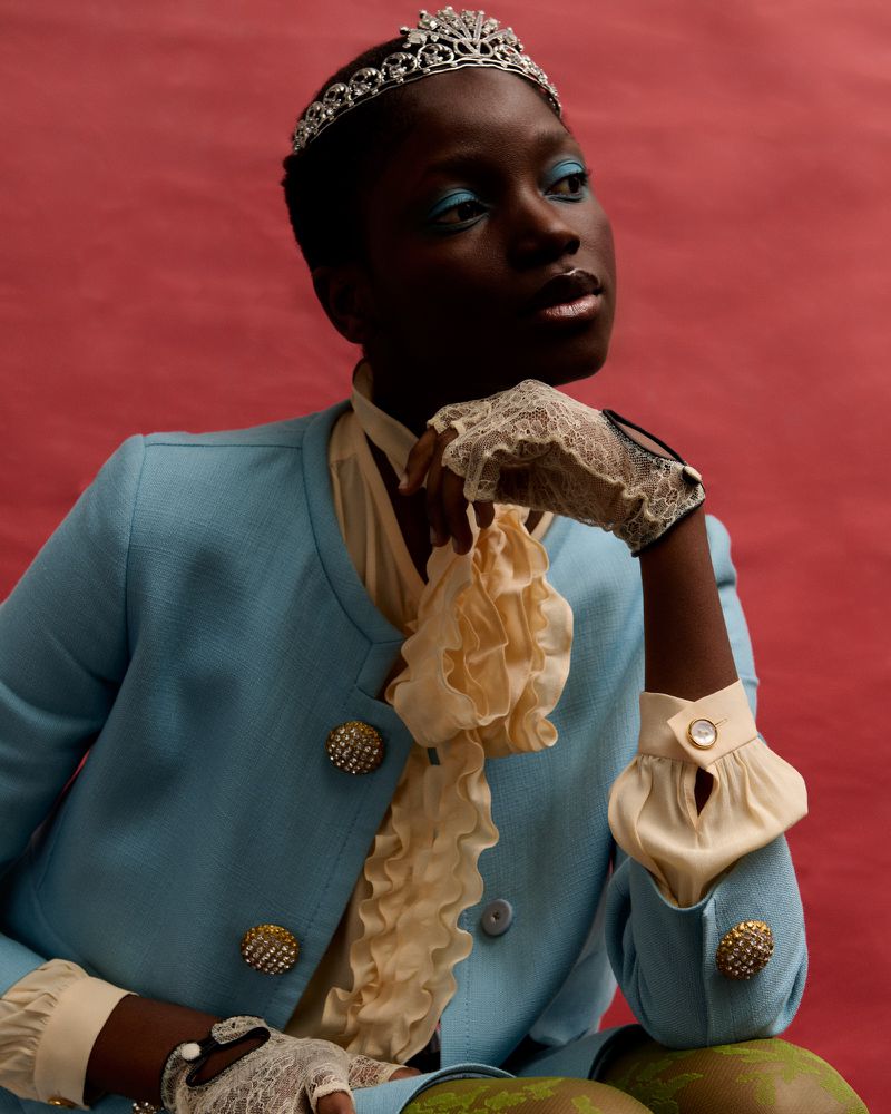Headshot of Black woman with tiara, light blue jacket, lace gloves, and bright blue eyeshadow against red backdrop in studio lighting.