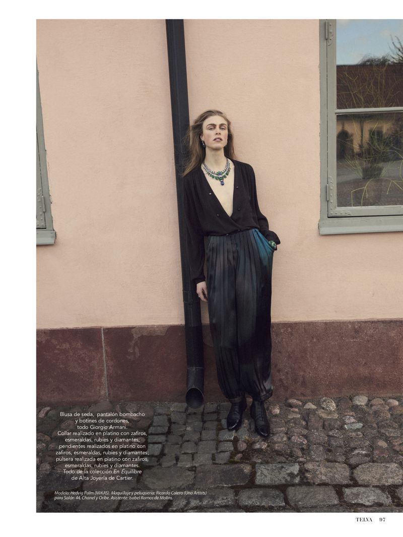 A blonde woman models a dark silk blouse and satin pants with jewelry while leaning against a pink wall on a cobblestone street.