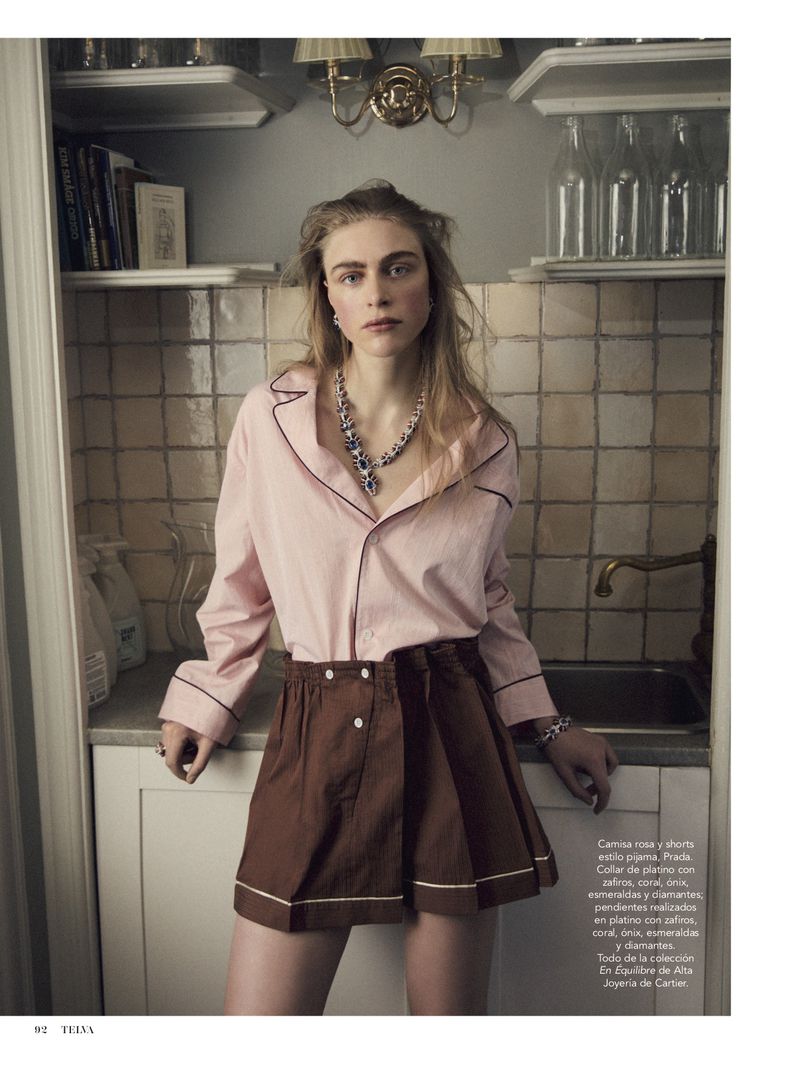 Blonde woman posing in a pink pajama top and brown shorts in an indoor setting; she is wearing a blue statement necklace and bracelet.